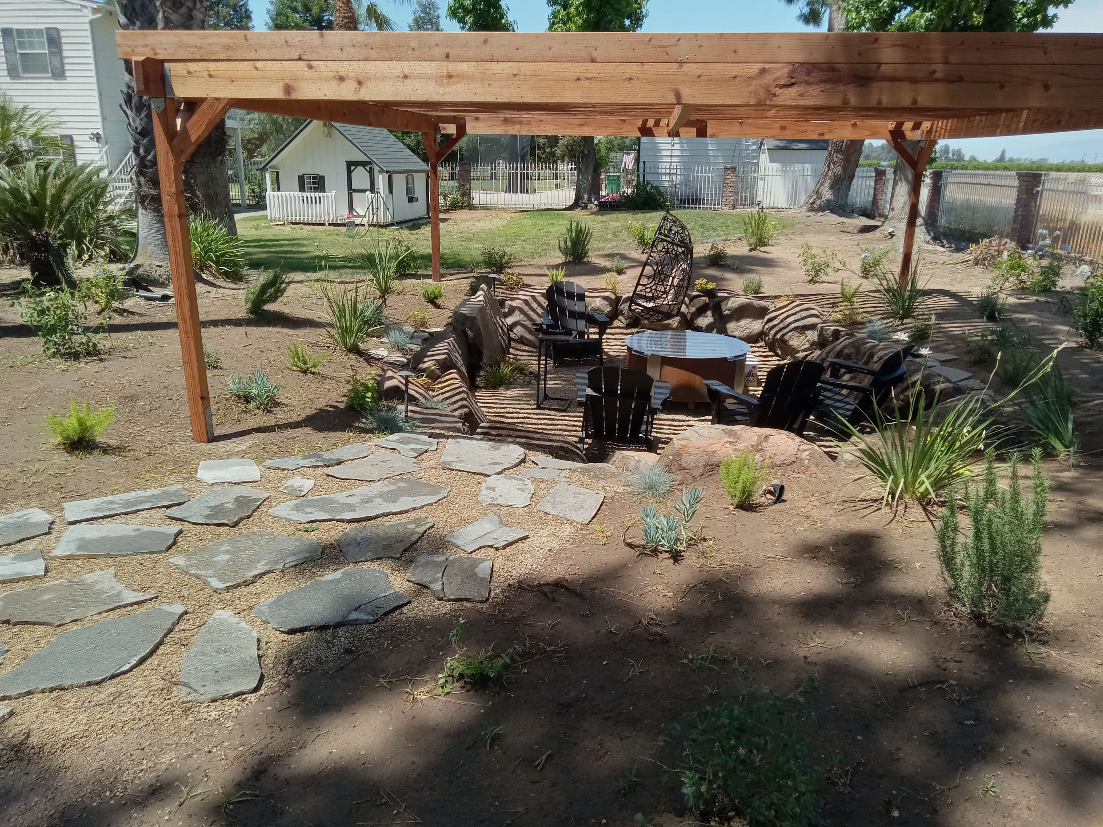 Cozy outdoor seating area with stone path and shaded pergola in a landscaped garden.