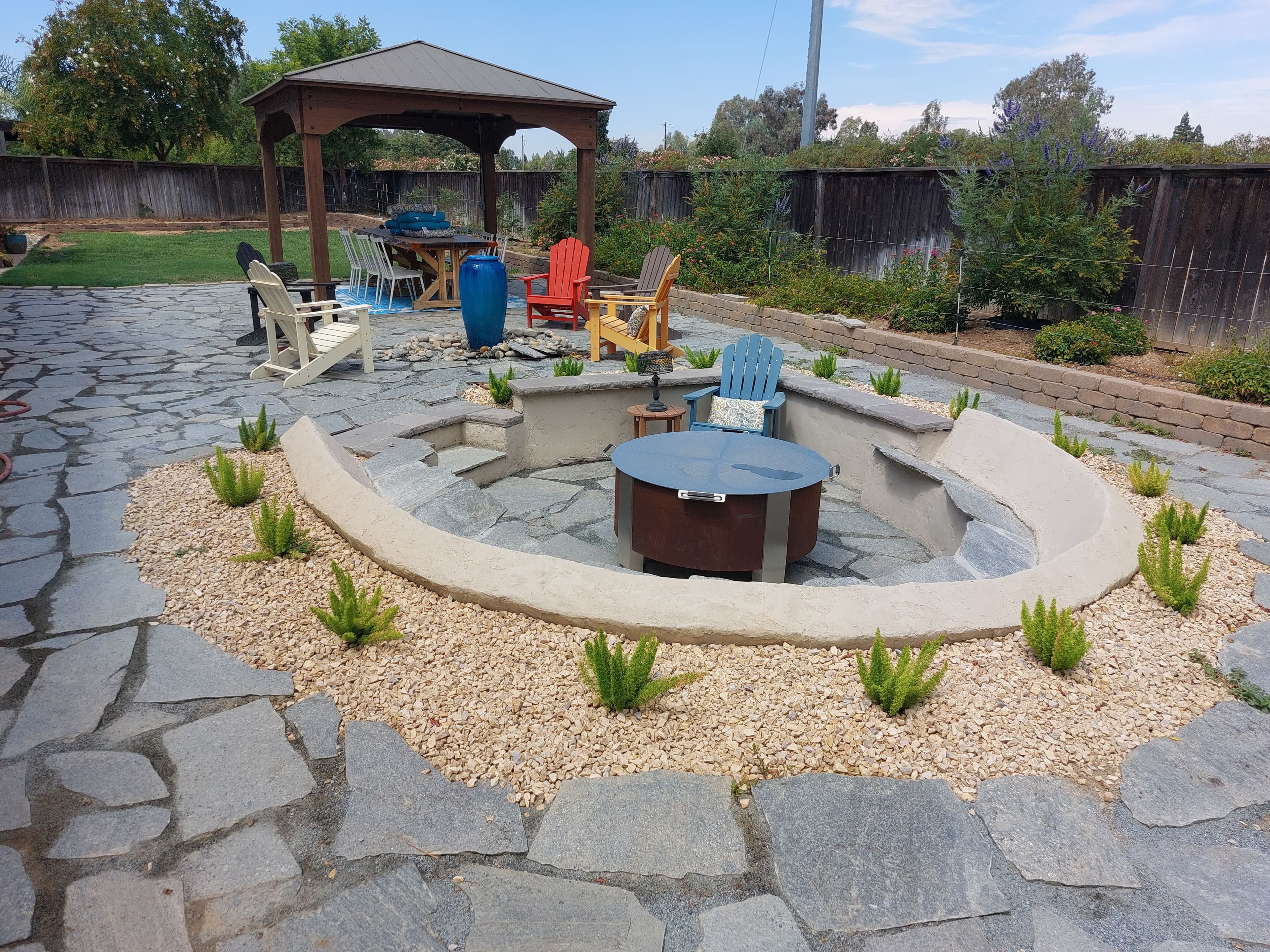 Outdoor patio with a fire pit, seating area, and gazebo surrounded by landscaping stones and plants.