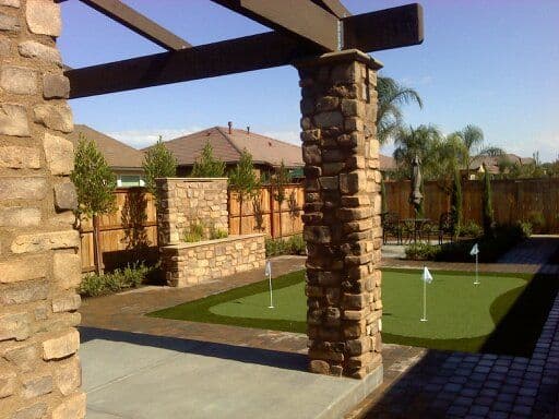 Backyard putting green with stone pillars and landscaped beds, featuring palm trees and fences.