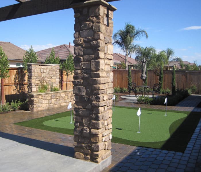 Backyard putting green with stone pillars, palm trees, and patio seating in a sunny landscape.