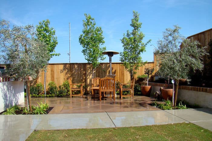 Outdoor patio with wooden furniture, lush greenery, and a heater under a blue sky.