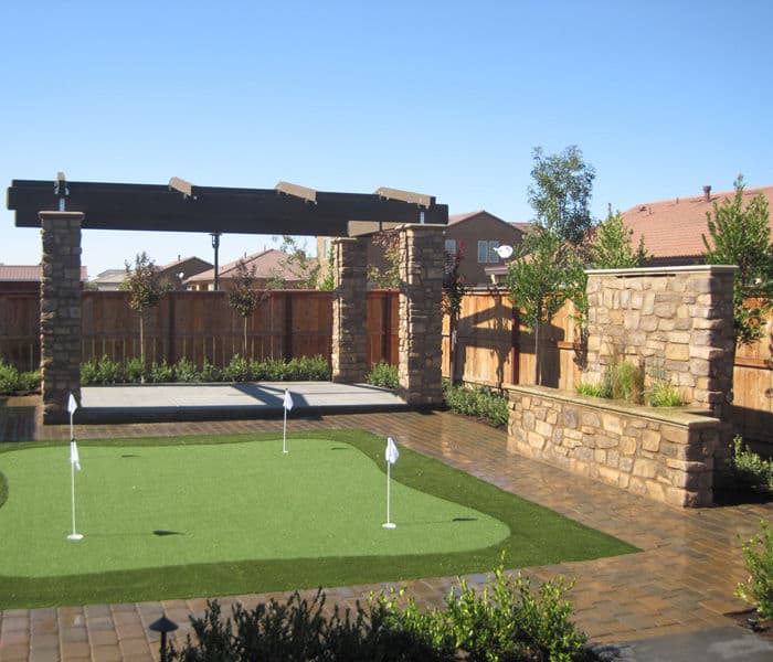 Backyard putting green with stone pillars and pergola, surrounded by greenery and landscaping.