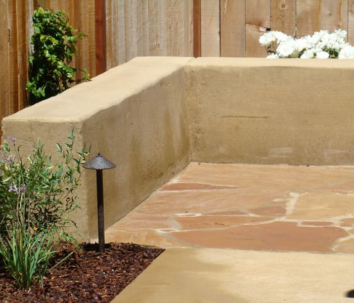 Garden corner with stone patio, low wall, and decorative plants near wooden fence.