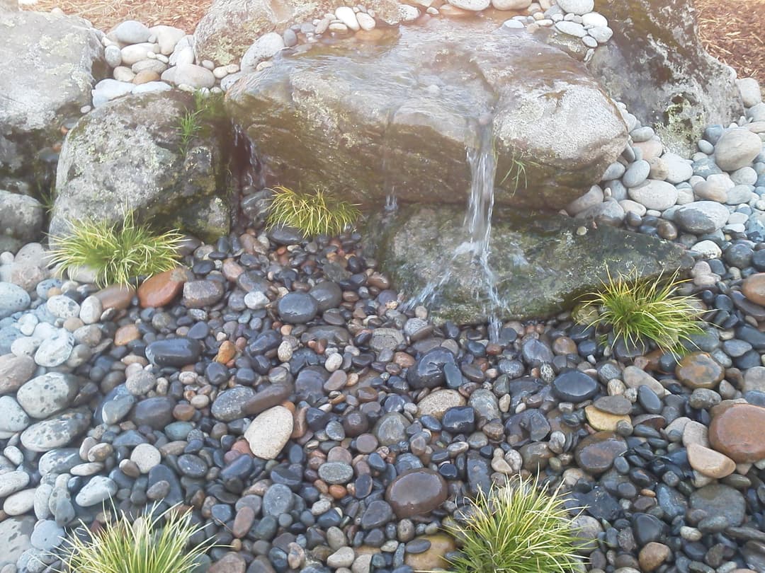 Flowing water over rocks in a tranquil garden with pebbles and grasses.