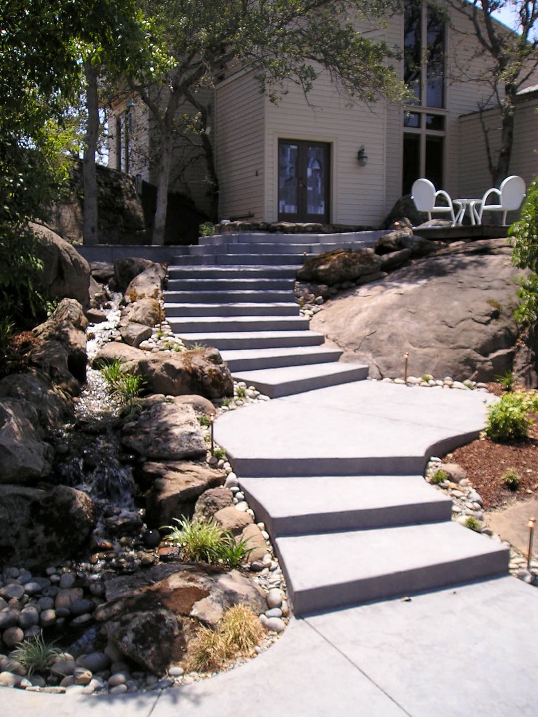 Concrete staircase with natural stone accents leading to a house, surrounded by greenery and water feature.