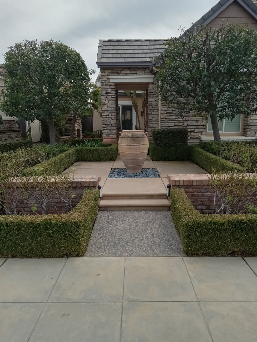 Front yard landscaping featuring a stone house, trimmed hedges, and a decorative clay pot fountain.