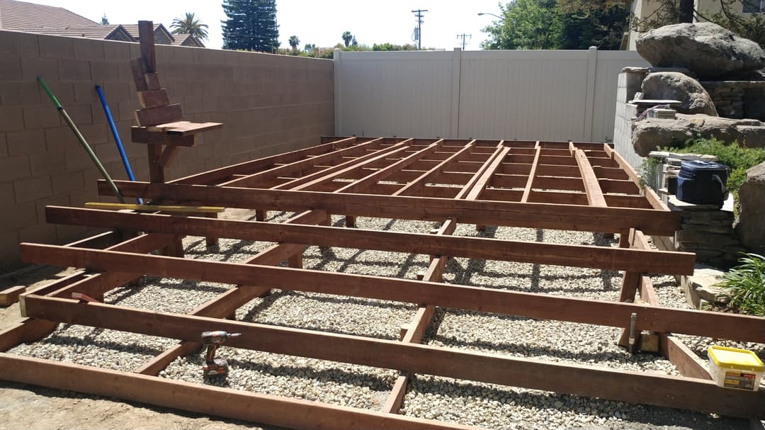 Wooden deck frame construction on gravel base in backyard, tools and materials visible.