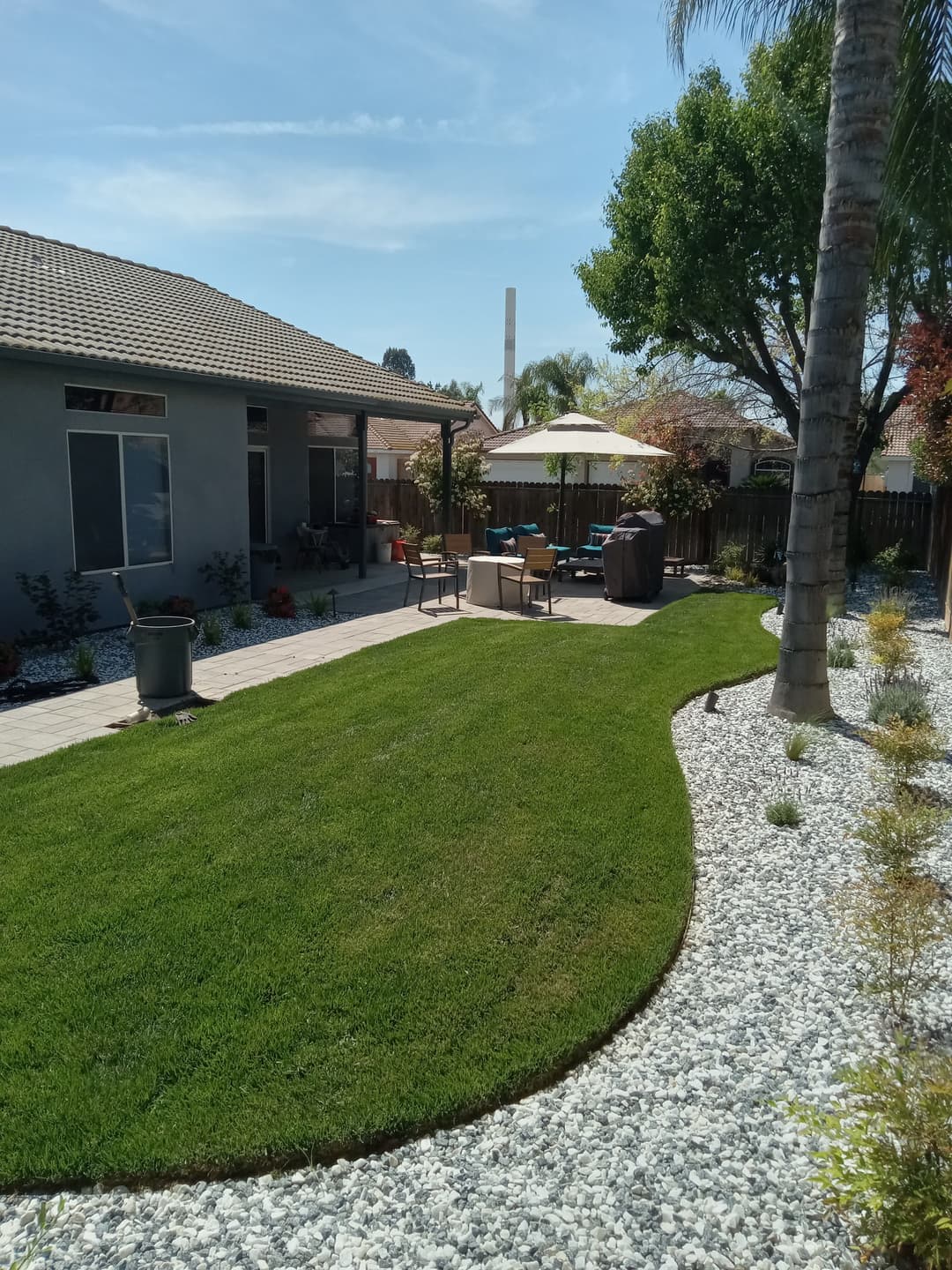 Lush green backyard with seating area, patio umbrella, and landscaped gravel beds.