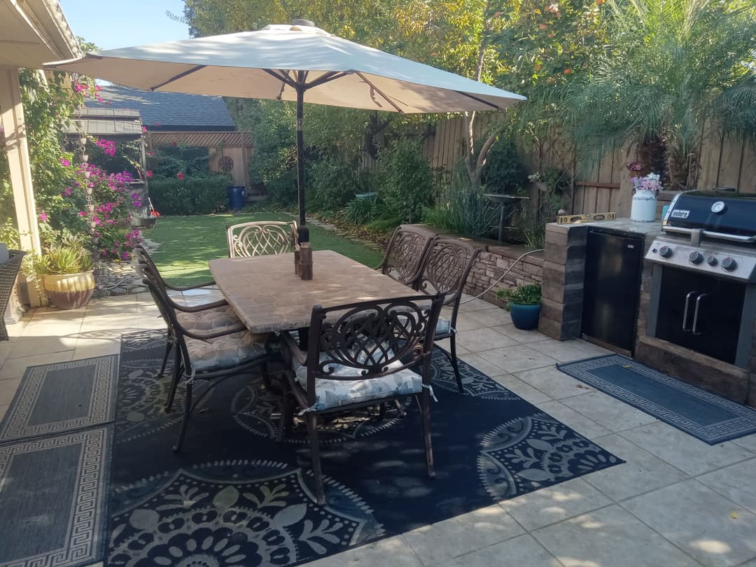 Outdoor patio with dining table, chairs, umbrella, grill, and lush garden in background.