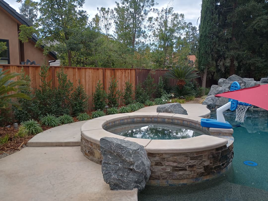 Backyard spa with natural stone, surrounded by greenery and a red umbrella structure.