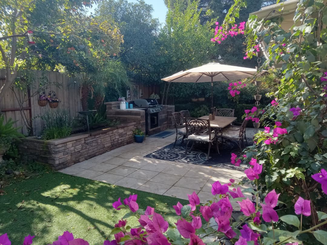 Lush backyard garden with flowers, dining area, and barbecue under a patio umbrella.