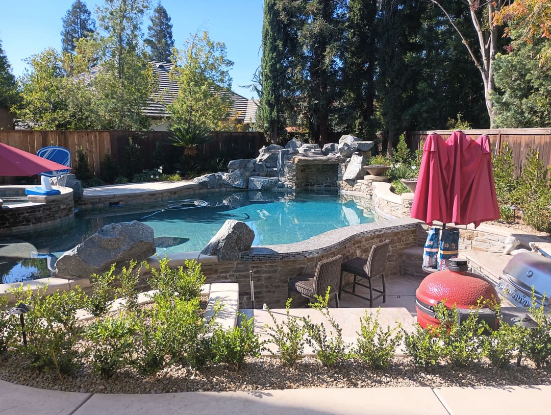 Luxury backyard pool with rocks, waterfall, and outdoor seating in a landscaped garden.