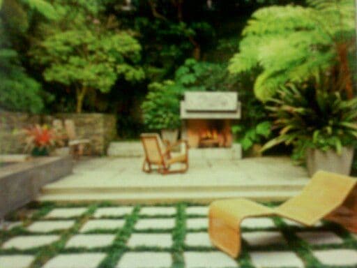 Lush garden patio with lounge chair, stone tiles, and grill surrounded by vibrant greenery.
