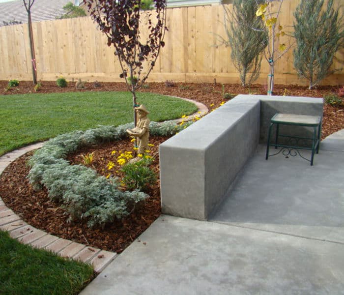 Concrete bench and decorative statue in a landscaped backyard with greenery and flower beds.