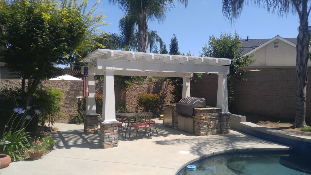 Backyard patio with a pergola, outdoor grill, and seating near a pool surrounded by greenery.
