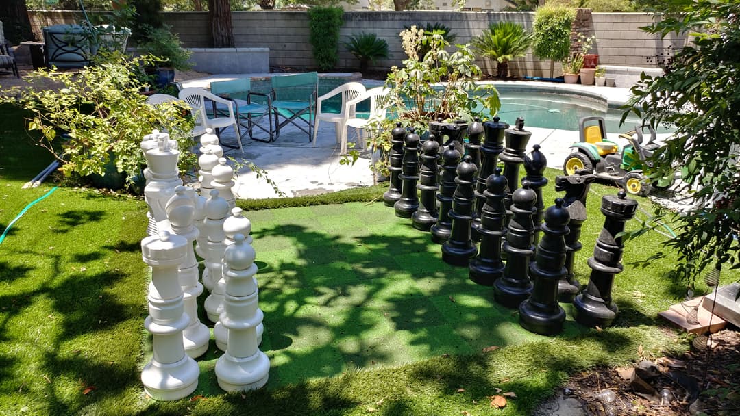 Giant outdoor chess set on green lawn beside a swimming pool with garden scenery.