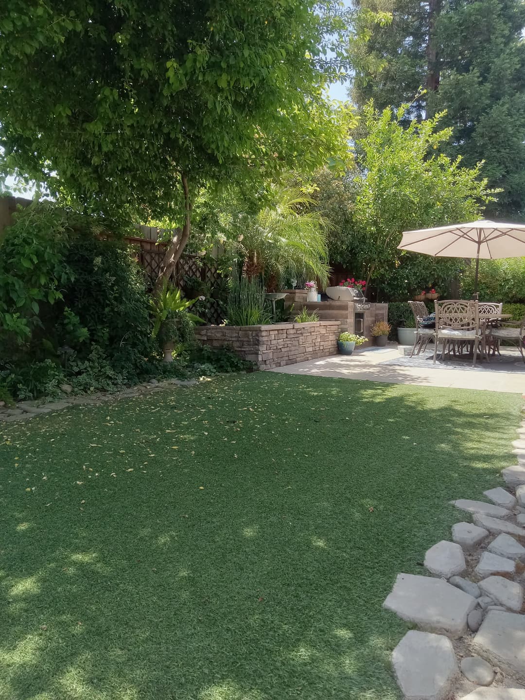 Lush backyard with artificial grass, stone pathway, garden plants, and patio seating area.