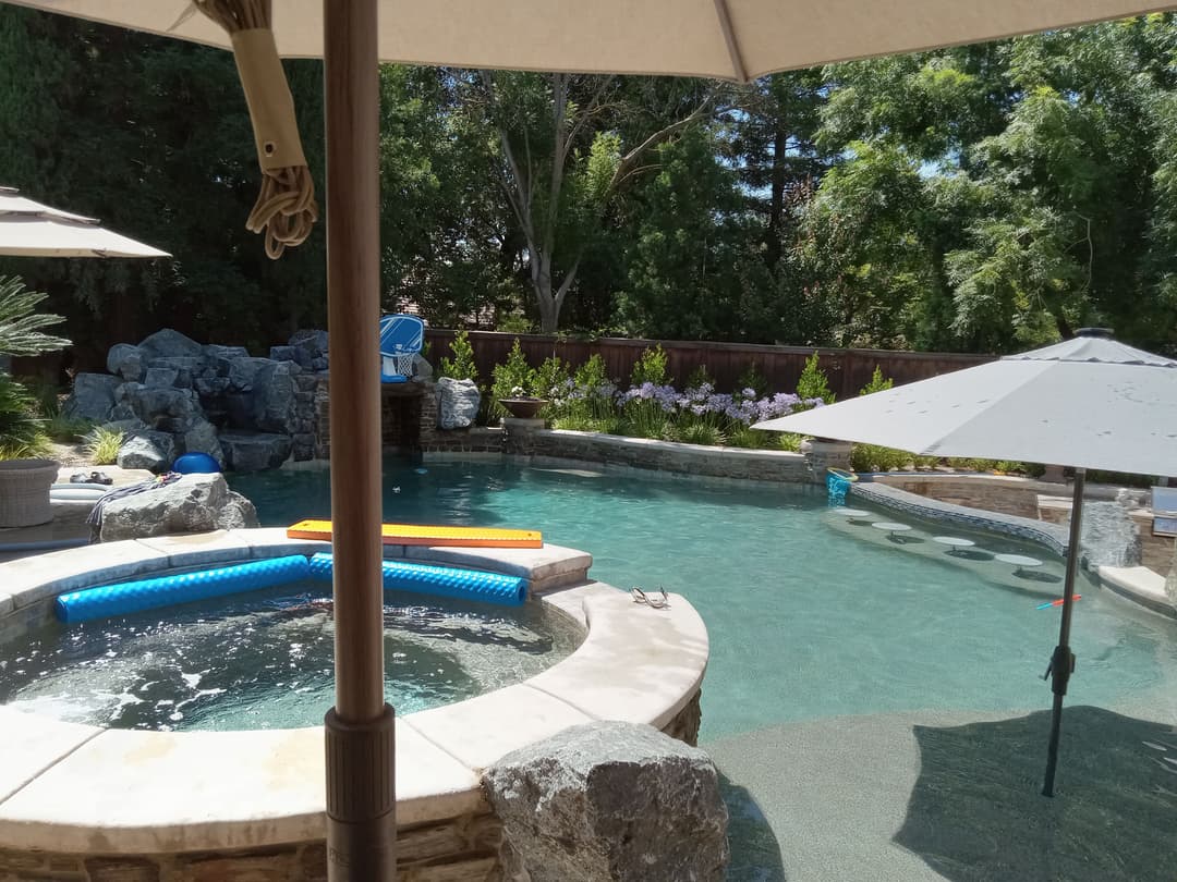 backyard oasis with a heated pool, spa, and shaded seating under umbrellas surrounded by greenery