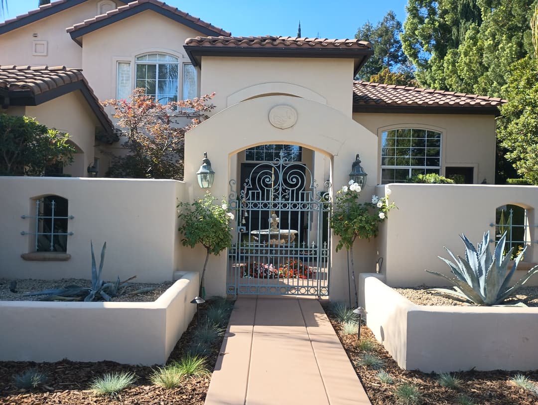 Charming stucco house with a decorative gate and lush landscaping in front.