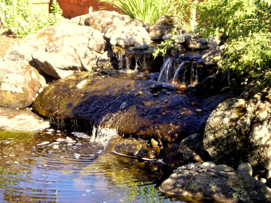 Serene garden pond with cascading waterfall over rocks and smooth pebbles.