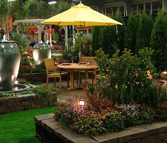 Cozy garden dining area with yellow umbrella, stone planters, and lush greenery at night.