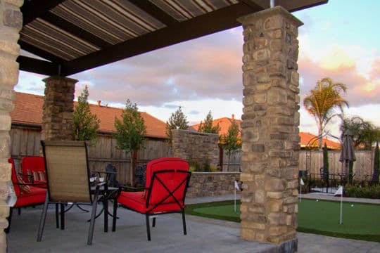 Patio area with red chairs, stone pillars, and putting green under a sunset sky.