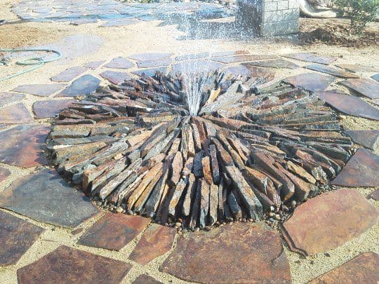 Rock fountain feature surrounded by stone pavement in a landscaped garden.
