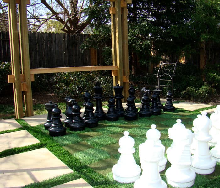 Giant outdoor chess set on grass with black and white pieces against a wooden backdrop.