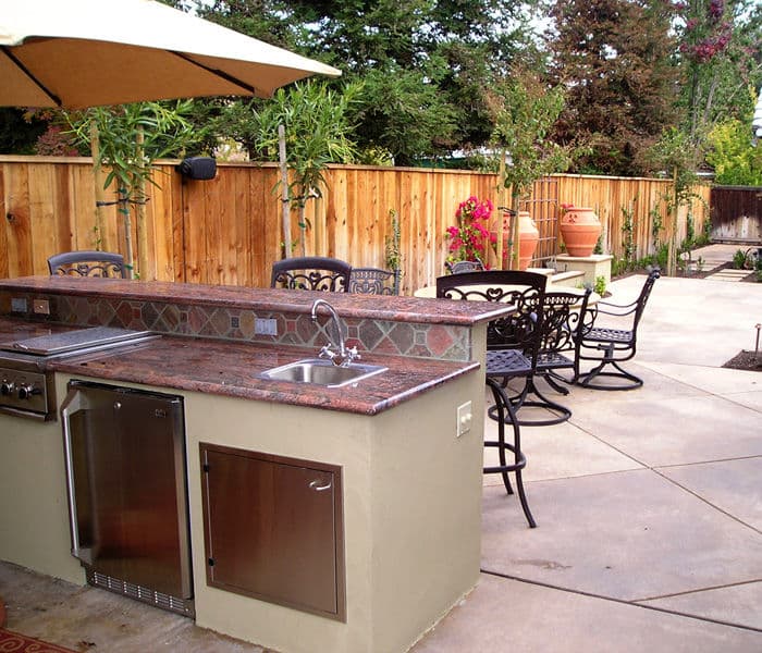 Outdoor kitchen with grill, sink, and patio seating surrounded by landscaping and wooden fence.