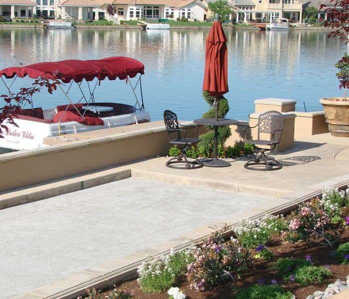 Lakeside patio with a table, chairs, umbrella, and colorful flower beds near a boat dock.