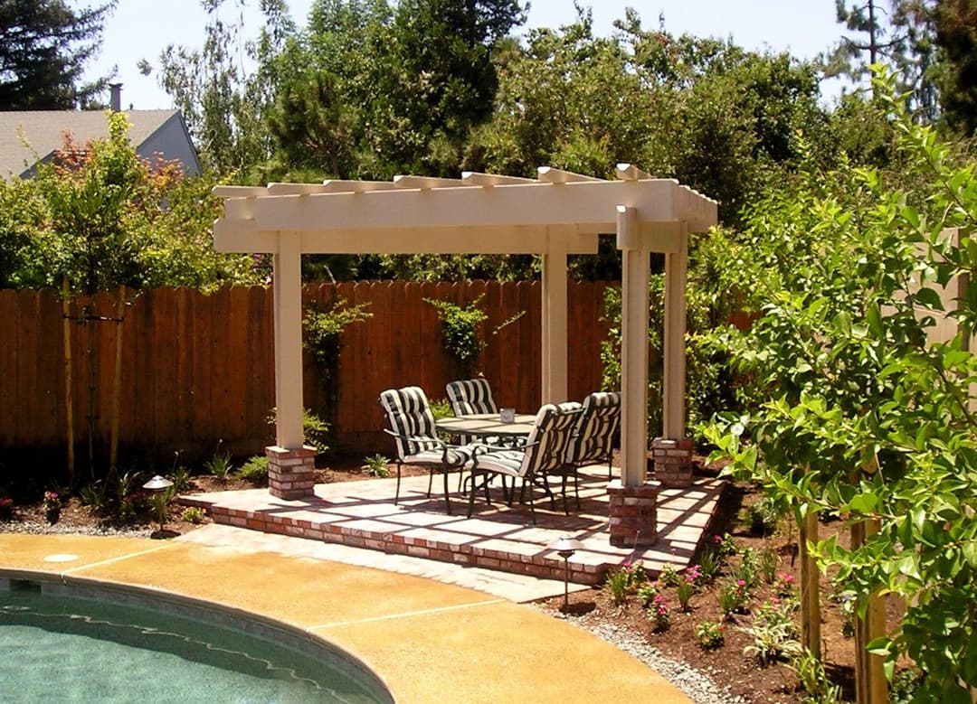 Outdoor pergola with dining set in landscaped backyard near a pool, surrounded by greenery.